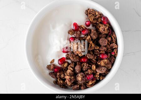 Schüssel mit griechischem Joghurt mit Schokoladenhafer und Granatapfelsamen auf einem weißen Marmortisch. Draufsicht. Gesundes Frühstück Stockfoto