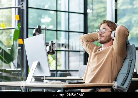Der junge, erfolgreiche Mann im Büro ruhte mit geschlossenen Augen und Händen hinter dem Kopf aus. Der Programmierer beendete seine Arbeit zufrieden mit den erzielten Ergebnissen und hörte Musik über Kopfhörer und Podcast online. Stockfoto