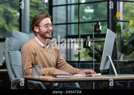 Junger erfolgreicher Unternehmer, der im Büro mit einem Computer arbeitet, Mann in legerer Kleidung, der lächelt und über die Tastatur tippt, Geschäftsmann, der mit seinen Leistungen und seinem Einkommen zufrieden ist. Stockfoto