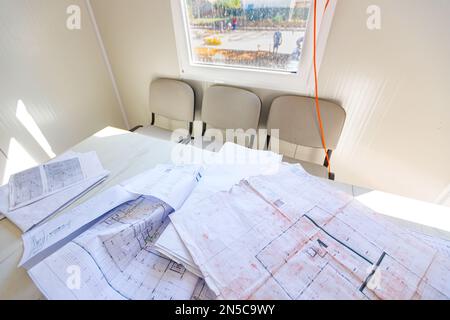 Masons Schreibtisch in einem Bürocontainer mit einem Haufen unordentlicher Architekturzeichnungen, Papierlayouts, Plänen. Fenster mit Blick auf die Baustelle Stockfoto