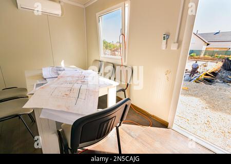 Masons Schreibtisch in einem Bürocontainer mit einem Haufen unordentlicher Architekturzeichnungen, Papierlayouts, Plänen. Fenster mit Blick auf die Baustelle Stockfoto