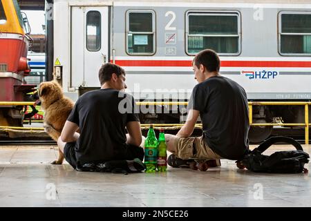 Zwei junge Männer, die sich unterhalten, mit ihrem Hund reisen, sitzen auf dem Bahnsteig Glavni Kolodvor. Zagreb, Kroatien, Europa, Europäische Union Stockfoto