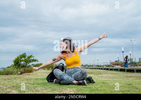 Fröhliche junge lateinin argentinischer ethnischer Herkunft, gekleidet in gelber und weißer Haut, mit offenen Armen, die auf dem Gras sitzen und lächeln, mit bewölktem Himmel i. Stockfoto