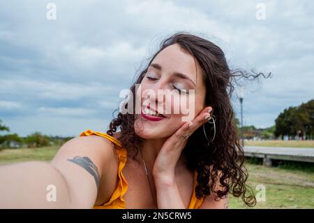Selfie-Porträt einer jungen süßen argentinischen Latina-Frau, weiß mit roten Lippen und gelber Kleidung, sie ist mit geschlossenen Augen und lächelt mit ihr Stockfoto