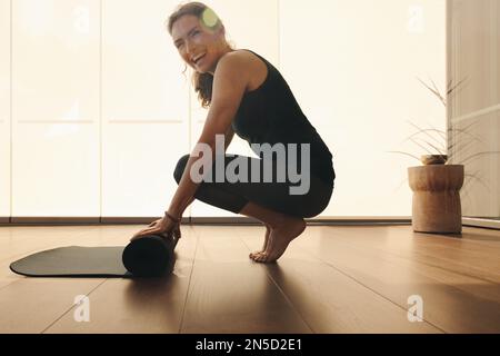 Reife Frau lächelt in die Kamera, während sie nach einer Yoga-Session eine Trainingsmatte hochklappte. Glückliche Frau, die zu Hause eine gesunde Trainingsroutine verfolgt. Sen Stockfoto
