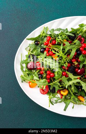 Sommer köstlicher Salat mit Rucola und frischen Beeren, roten und schwarzen Johannisbeeren, süßen Kirschen und schwarzen Maulbeeren, Salat auf einem ovalen weißen Teller Stockfoto
