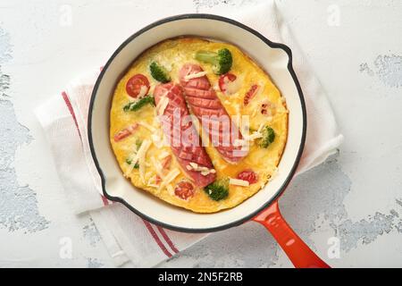 Omelette oder Frittata mit zwei Würstchen, geschmolzenem Käse, Kirschtomaten, Brokkoli und Petersilie auf roter Pfanne oder Eisentopf auf weißem, altem Betonrücken Stockfoto