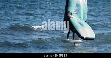 Ein Mann trägt Flügelfolien mit handgeführten aufblasbaren Flügeln und Tragflächensurfbrettern in einem blauen Ozean, reitet auf einem Windflügelbrett, surft auf den Wellen Stockfoto
