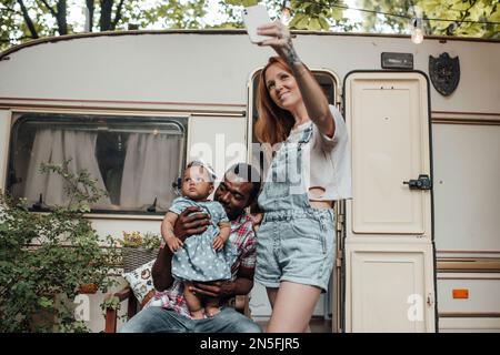 Gemischte Familien mit dunkel gehäuteter Kleinkindertochter vor dem Wohnwagen machen Fotos für soziale Medien. Ein afroamerikanischer Mann, seine Frau und sein kleines Kind Stockfoto