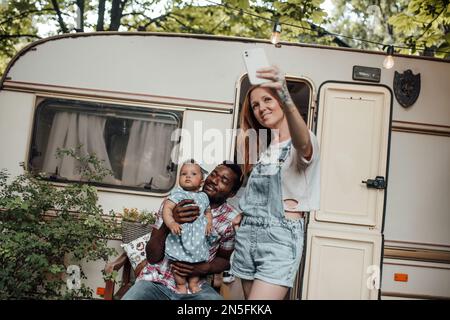 Gemischte Familien mit dunkel gehäuteter Kleinkindertochter vor dem Wohnwagen machen Fotos für soziale Medien. Ein afroamerikanischer Mann, seine Frau und sein kleines Kind Stockfoto