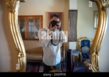 Ein junger weißer Fotograf posiert für ein Selfie mit einer professionellen Kamera in einem Golden Frame Spiegel in einem schicken Old School Wohnzimmer Stockfoto