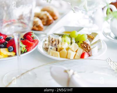 Tisch für Bankett serviert. Teller mit Obst und Vorspeisen, transparente, moderne, stilvolle Weingläser und Besteck auf weißem Tischtuch. Stockfoto