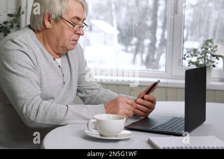 Ein gut aussehender, seriöser Seniorenmann, der eine Brille trägt und gleichzeitig mit seinem Laptop am Schreibtisch sitzt. Pensionierter Mann, der von zu Hause aus arbeitet Stockfoto
