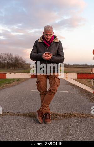 Mann mittleren Alters in Winterkleidung mit Smartphone auf einer Landstraße - das Konzept des Menschen in der Freizeit Stockfoto