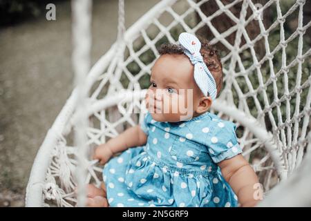 Ein kleines Mädchen in blauem, gepunktetem Kleid hat positive Emotionen, sitzt auf weißem, makramfarbenen Baumwollseil, Kokon hängt. afroamerikanisches Baby-Rumpf Stockfoto