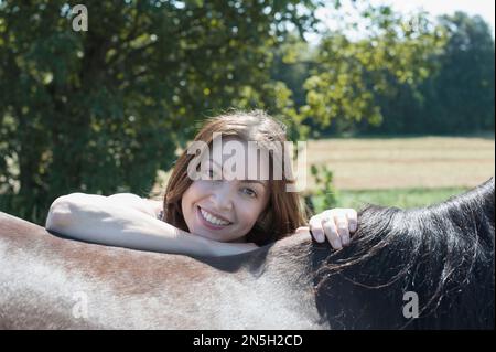 Porträt einer erwachsenen Frau, die sich auf den Rücken eines braunen Pferdes lehnt und lächelt, Bayern, Deutschland Stockfoto