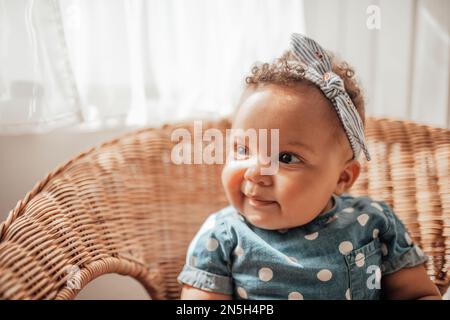 Ein kleines Mädchen in blauem Punktkleid hat positive Emotionen, wenn es auf einem Stock sitzt, hell und im Gesicht schattiert. afroamerikanisches Baby entspannt A Stockfoto