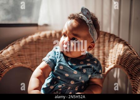 Ein kleines Mädchen in blauem Punktkleid hat positive Emotionen, wenn es auf einem Stock sitzt, hell und im Gesicht schattiert. afroamerikanisches Baby entspannt A Stockfoto