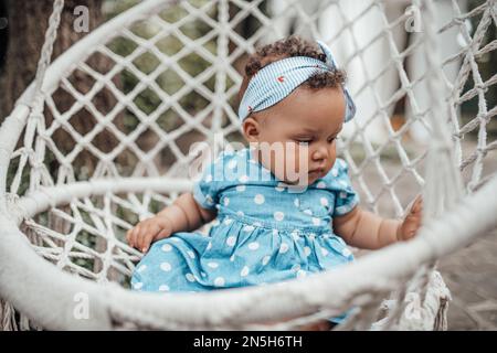 Ein kleines Mädchen in blauem, gepunktetem Kleid hat positive Emotionen, sitzt auf weißem, makramfarbenen Baumwollseil, Kokon hängt. afroamerikanisches Baby-Rumpf Stockfoto