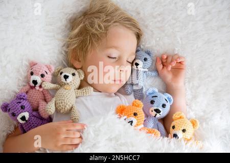 Kleines Kleinkind, niedlicher blonder Junge, der mit vielen Teddybären schläft, handgemachtes Amigurumi-Spielzeug zu Hause im Bett Stockfoto