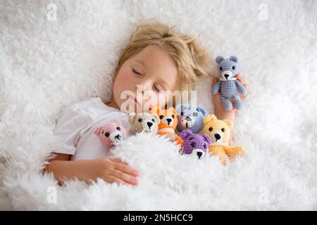 Kleines Kleinkind, niedlicher blonder Junge, der mit vielen Teddybären schläft, handgemachtes Amigurumi-Spielzeug zu Hause im Bett Stockfoto