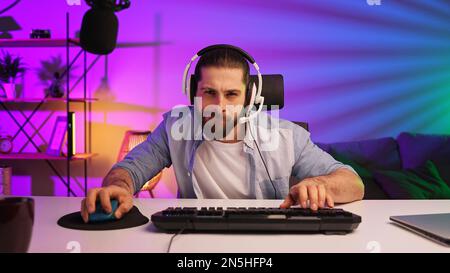 Ein bärtiger Gamer in einem Gaming-Headset gewinnt ein Videospiel. Cybersportler in einem Neonraum. Lifestyle-Gamer, Online-Spiele. Sieg im E-Sport Stockfoto