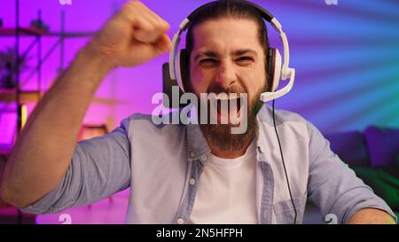 Ein bärtiger Gamer in einem Gaming-Headset gewinnt ein Videospiel. Cybersportler in einem Neonraum. Lifestyle-Gamer, Online-Spiele. Sieg im E-Sport Stockfoto