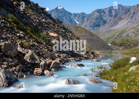 Alpenfluss Karakabak fließt im Sommer die Steine im Altai-Gebirge hinunter mit einer bewegenden Silhouette eines Mannes an der Küste. Stockfoto