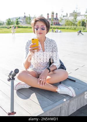 Eine Frau mit farbenfroher Sonnenbrille sitzt auf einer offenen Holzbühne im öffentlichen Park. Hübsche Frau in legerer Kleidung, die nach dem Motorroller SMS auf dem Smartphone schreibt Stockfoto