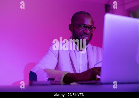 Ein Schwarzer in einem weißen Anzug und einer Brille arbeitet an einem Laptop. Ein Mann mit Brille arbeitet abends im Büro Stockfoto