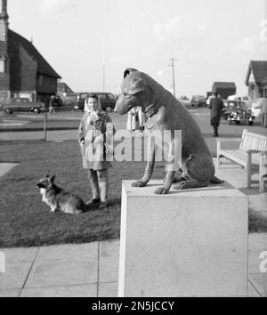 1963, historisch, eine Dame mit ihrem kleinen Hund neben ihr, stehend, auf eine Statue eines Hundes, 'Snooks' auf dem Marktplatz, Aldeburgh, Suffolk, England, Großbritannien. Die Bronzestatue wurde 1961 zum Gedenken an den örtlichen Aldeburgh GP Dr. Robin Acheson errichtet, als sein Hund Snooks, der dem Arzt folgte, während er seine Anrufe machte, zu einem vertrauten Anblick rund um die Küstenstadt wurde. Stockfoto