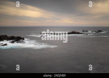 Sonnenuntergang am Point Lobos State Natural Reserve, lange Exposition, mit dem Ozean und Felsen vor einem teilweise bewölkten Himmel mit Orangentönen. Carmel, Califo Stockfoto