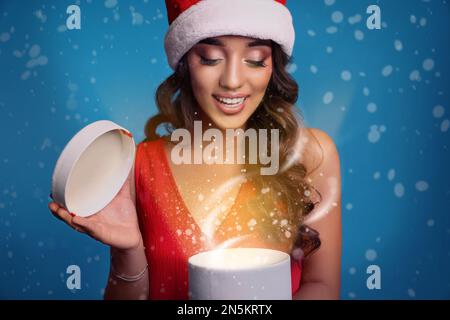 Schöne Frau mit Weihnachtsmann-Hut, die ein Weihnachtsgeschenk auf blauem Hintergrund eröffnet Stockfoto