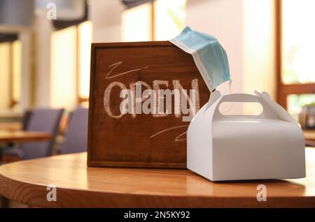 OFFENES Schild, gepackte Bestellung zum Mitnehmen und Schutzmaske auf dem Tisch im Restaurant. Gastronomie während der Coronavirus-Quarantäne Stockfoto