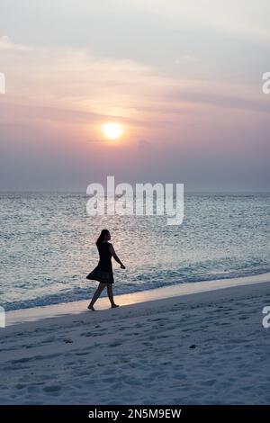 Eine Frau, die alleine am Sandstrand bei Sonnenuntergang am Ufer spaziert, in Silhouette, Rückblick, Malediven, Beispiel für Alleinreisen. Stockfoto