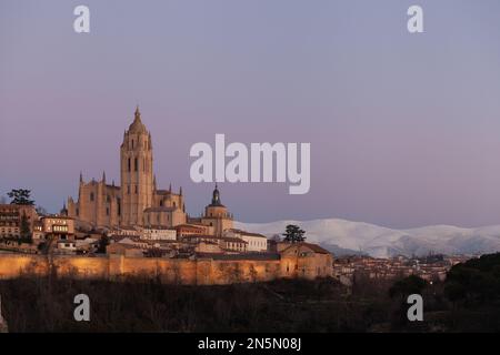 Segovia, Spanien - 4. Januar 2022: Panoramablick auf Segovia mit den Türmen der Kathedrale bei Sonnenuntergang und violettem Himmel im Winter mit dem Guadarrama-Gebirge Stockfoto