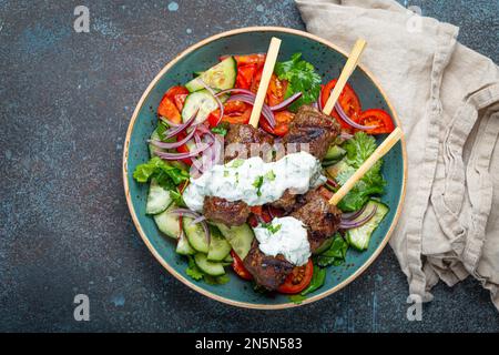 Gegrillte Spießfleisch-Rindfleisch-Kebabs auf Stöcken serviert mit frischem Gemüsesalat auf einem Teller auf rustikalem Betonhintergrund von oben. Traditioneller Mittelteil Stockfoto