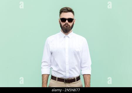 Porträt eines gutaussehenden, bärtigen Geschäftsmanns mit weißem Hemd und schwarzer Sonnenbrille, der mit ernstem und selbstbewusstem Gesichtsausdruck steht. Studioaufnahme im Innenbereich isoliert auf hellgrünem Hintergrund. Stockfoto