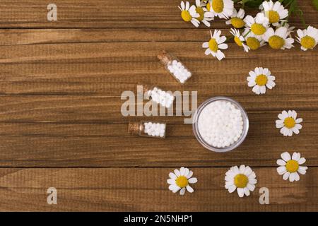 Homöopathische Globula und Glasflasche auf natürlichem Holzhintergrund neben medizinischen Kamillenblüten. Alternative Homöopathie Medizin Kräuter, Heilung Stockfoto