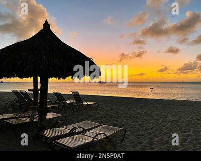 Blick über einen Strand in Aruba und den Sonnenuntergang über dem Karibischen Meer. Sonnenschirm und Stühle am Strand, Segelboot am Horizont. Stockfoto