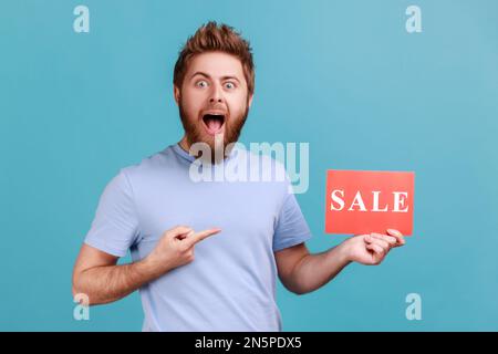 Portrait eines aufgeregten, bärtigen Mannes, der mit dem Finger auf die Karte zeigt, mit einem Verkaufswort, der den Mund in Erstaunen öffnet und schockiert von Rabatten aussieht. Innenstudio, isoliert auf blauem Hintergrund. Stockfoto