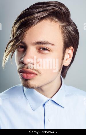 Porträt eines jungen, gutaussehenden Mannes mit langen Fransen auf den Augen, der mit mysteriösem Blick in die Kamera schaut, ein blaues Hemd trägt. Studioaufnahmen im Innenbereich isoliert auf grauem Hintergrund. Stockfoto