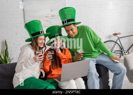 Glückliche Frauen und ein afroamerikanischer Mann in grünen Hüten, der ein Glas dunkles Bier hält, während er auf den Laptop schaut Stockfoto