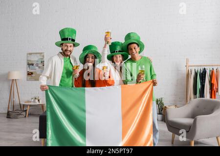 Fröhliche und multikulturelle Freunde in grünen Hüten mit Gläsern Bier und irischer Flagge, während der St. Patrick Day gefeiert wird Stockfoto