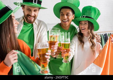 Fröhliche und gemischtrassige Freunde in grünen Hüten mit Gläsern Bier und irischer Flagge, während der St. Patrick Day gefeiert wird Stockfoto