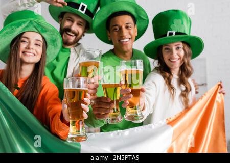 Fröhliche und multikulturelle Freunde in grünen Hüten mit Gläsern Bier und irischer Flagge während des St. Patrick Day Stockfoto