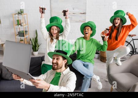 Ein bärtiger Mann hielt ein Notebook in der Nähe glücklicher multiethnischer Freunde mit Gläsern dunkles Bier, während er den St. Patrick Day feierte Stockfoto