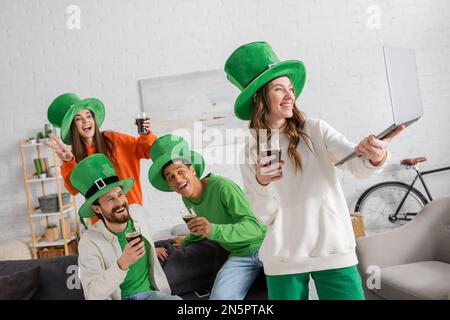 Fröhliche Frau, die ihr Notebook in der Nähe glücklicher multiethnischer Freunde hält, mit Gläsern dunkles Bier, während sie den St. Patrick Day feiert Stockfoto