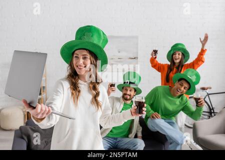 Lächelnde Frau, die ihr Notebook in der Nähe glücklicher multiethnischer Freunde hält, mit einem Glas dunklem Bier, während sie den St. Patrick Day feiert Stockfoto