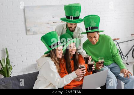 Fröhliche und multiethnische Freunde in grünen Hüten, die ein Glas dunkles Bier trinken, während sie auf ein Notebook schauen Stockfoto